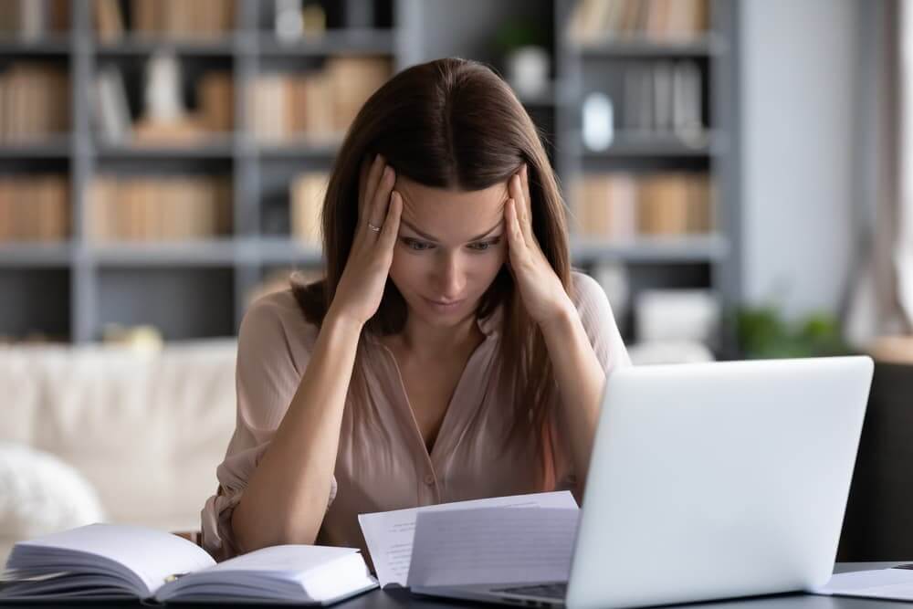 Woman with hands on temples looking down at laptop and papers.