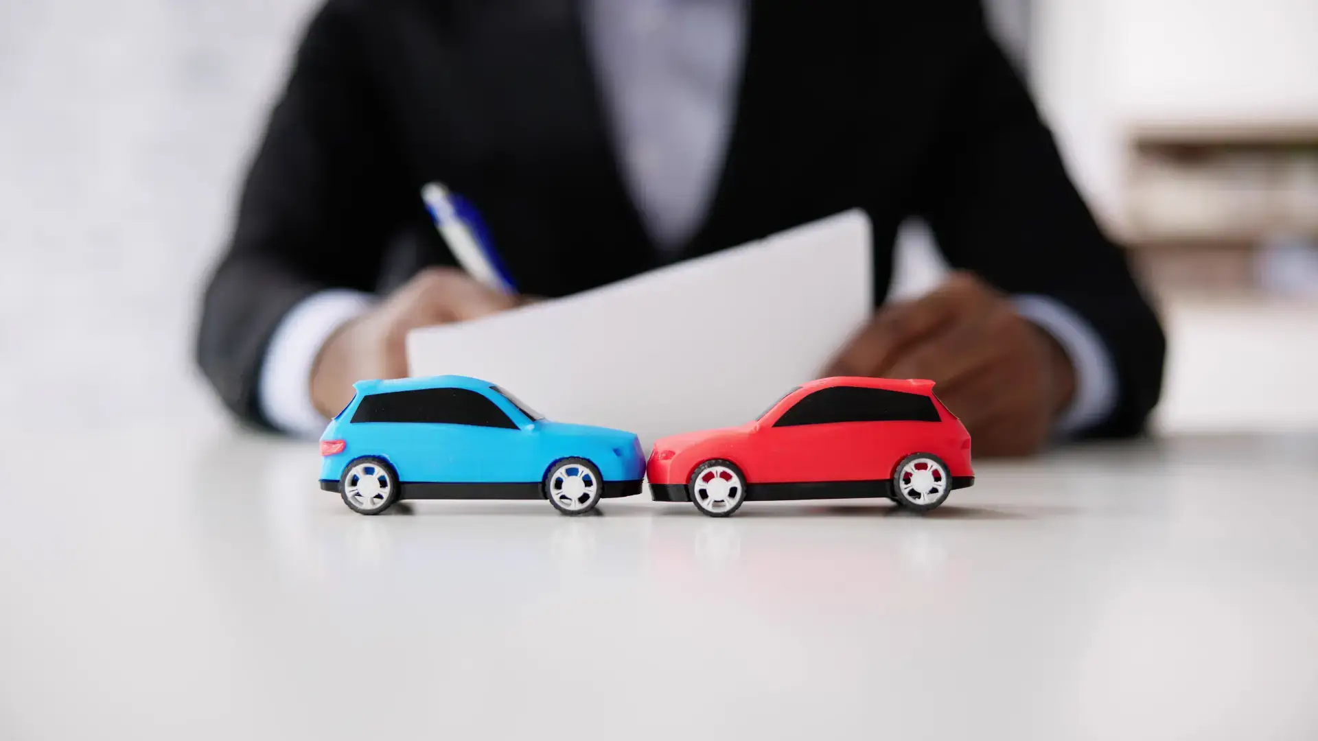 Two toy cars, one blue and one red, positioned as if in a collision, with a person in a suit writing in the background.
