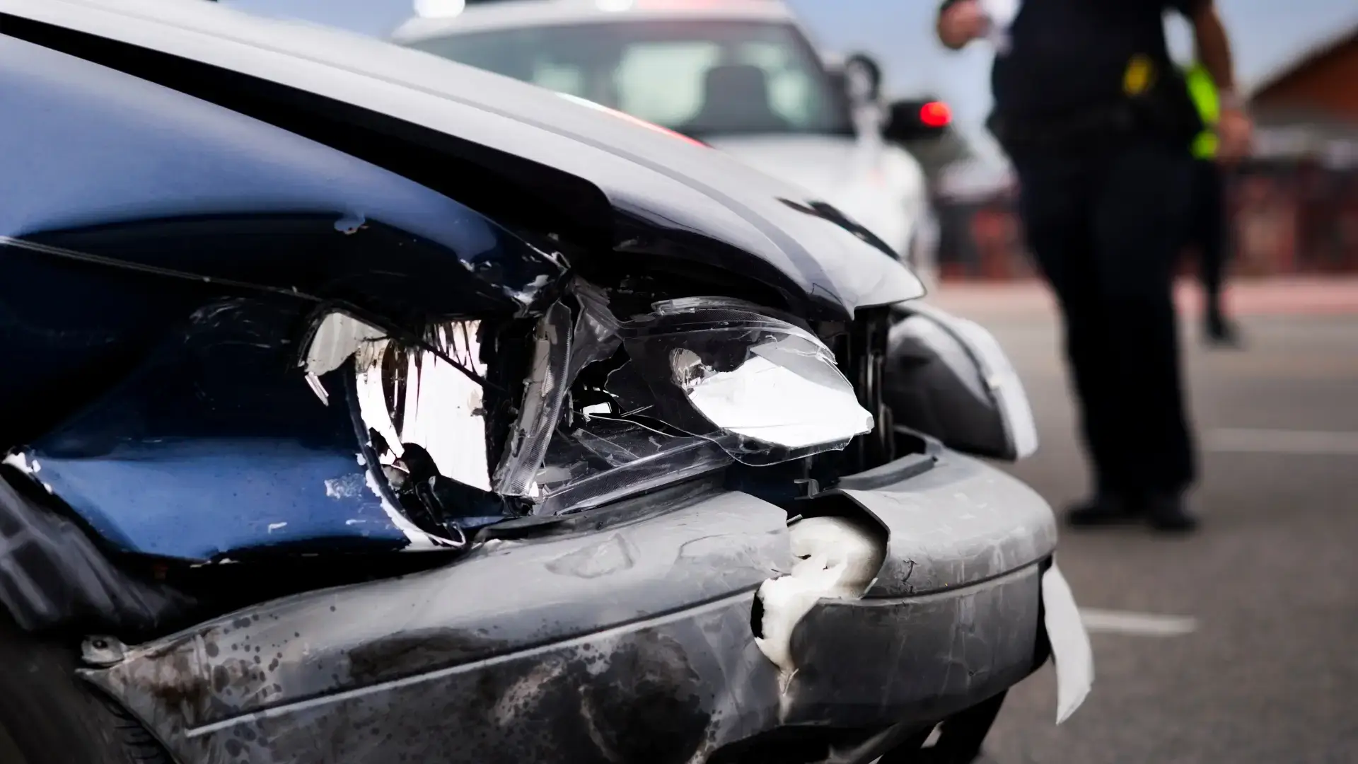 Close-up of a heavily damaged car front after an accident, with other vehicles and a person in the background.