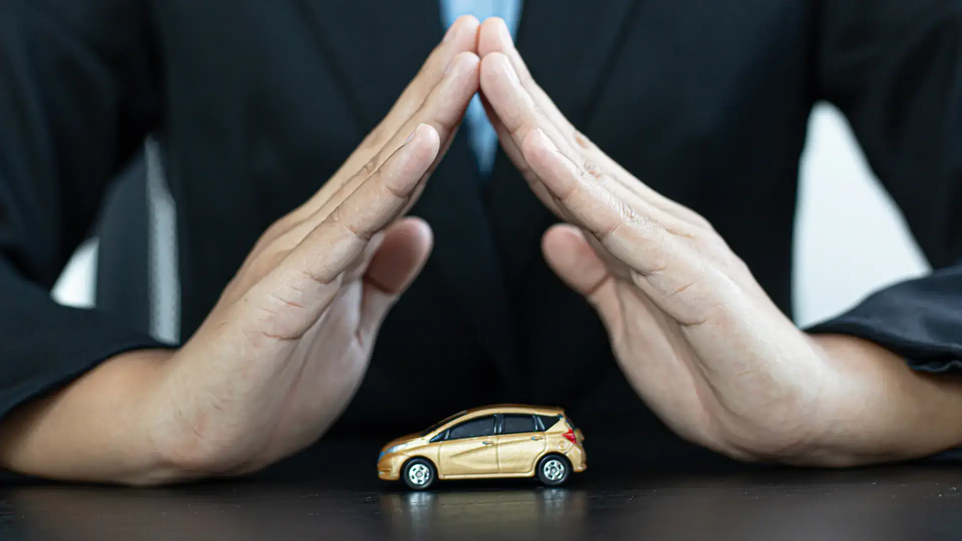 Hands forming a protective gesture over a small model car, symbolizing car insurance or protection.