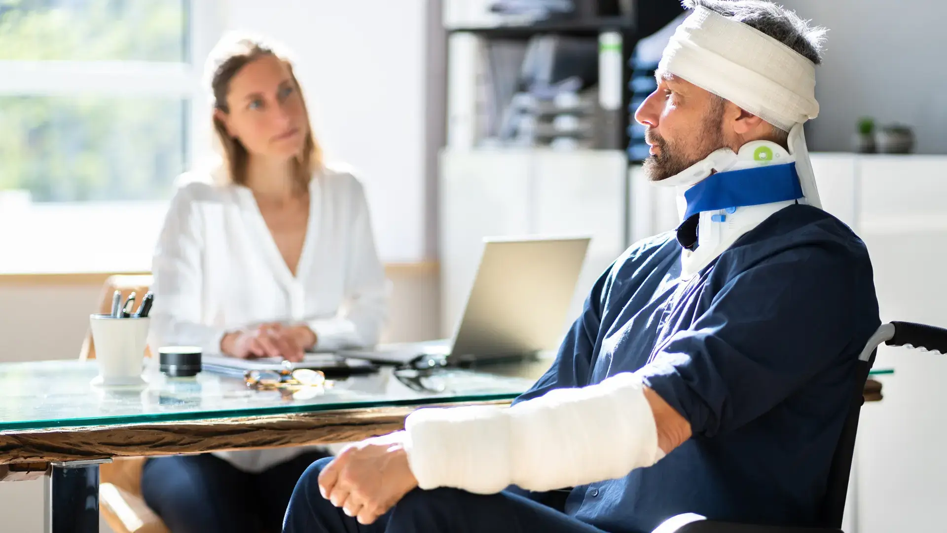 Injured person in a neck brace and arm bandages discussing with a healthcare professional at a desk.