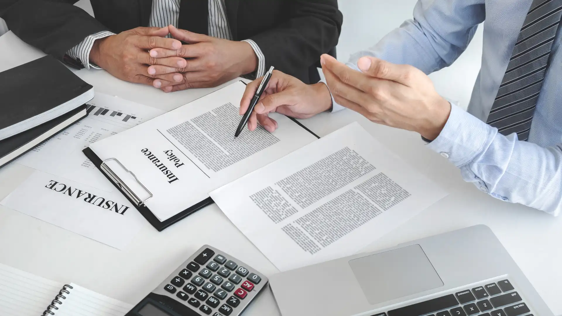 business meeting with two individuals discussing documents, one holding a pen to sign and the other gesturing while a calculator is placed on the table.