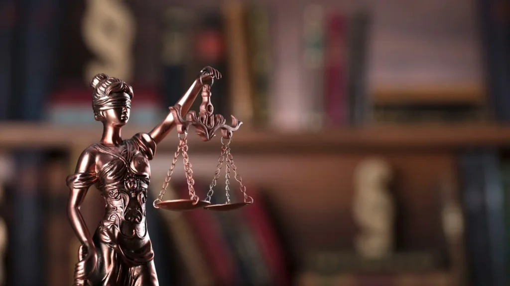 A close-up of a bronze Lady Justice figurine holding scales in a law office with blurred bookshelves in the background.