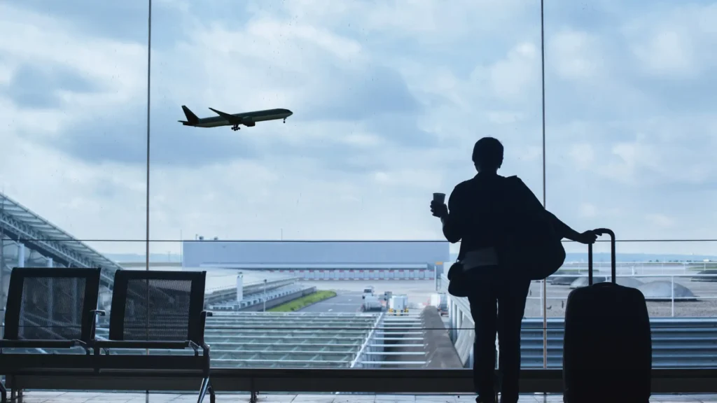 A person standing at the airport window, holding a cup and watching an airplane take off.