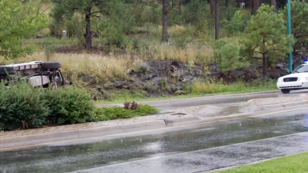 A wet road with a rainy ambiance, showing the reflection of the surrounding greenery and rocks.